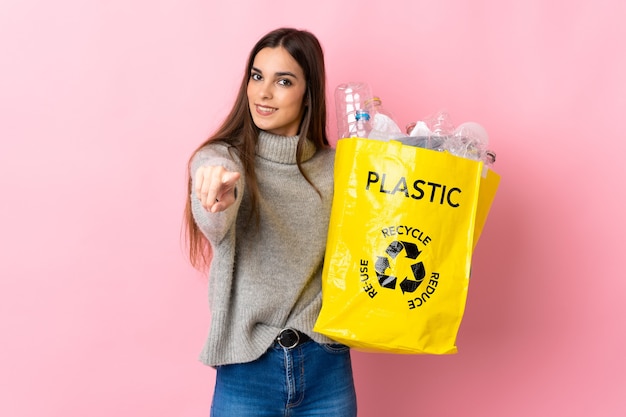 Jonge blanke vrouw met een zak vol plastic flessen om te recyclen geïsoleerd op roze achtergrond naar voren wijzend met gelukkige uitdrukking