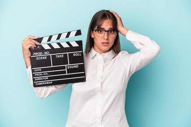 Foto jonge blanke vrouw met filmklapper geïsoleerd op blauwe achtergrond geschokt, ze heeft een belangrijke vergadering onthouden.