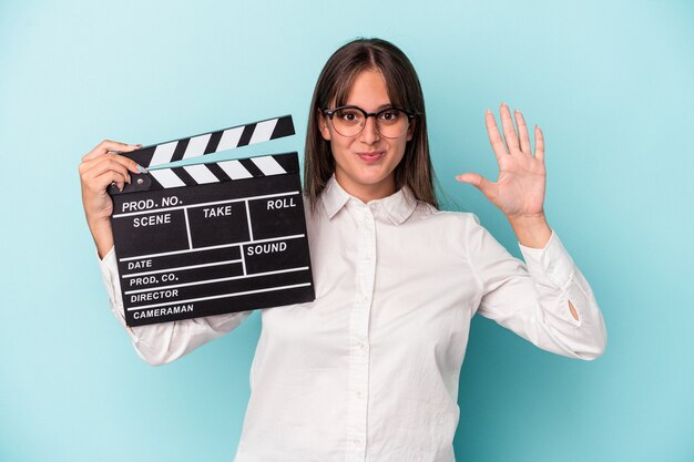 Jonge blanke vrouw met filmklapper geïsoleerd op blauwe achtergrond glimlachend vrolijk nummer vijf met vingers.