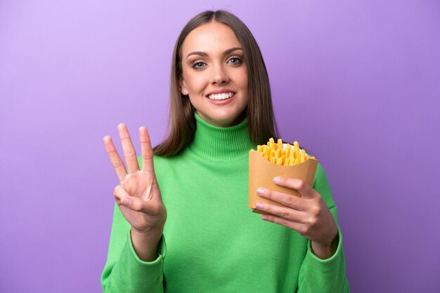 Jonge blanke vrouw met gefrituurde chips op paarse achtergrond gelukkig en drie tellen met vingers