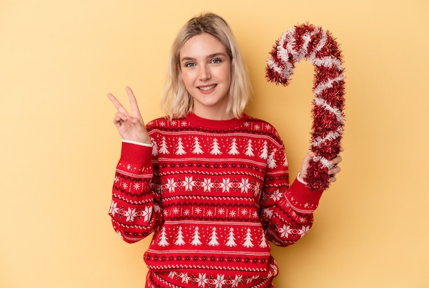 Jonge blanke vrouw met grote kerststok geïsoleerd op gele achtergrond vrolijk en zorgeloos met een vredessymbool met vingers.