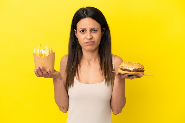 Jonge blanke vrouw met hamburger en patat geïsoleerd op gele achtergrond