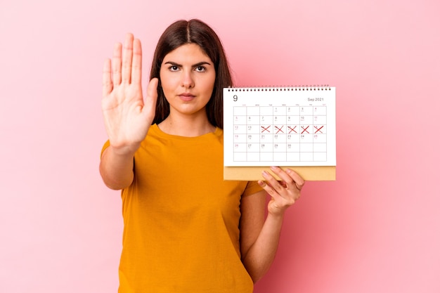 Jonge blanke vrouw met kalender geïsoleerd op een roze achtergrond die staat met uitgestrekte hand die een stopbord toont, waardoor je wordt voorkomen.