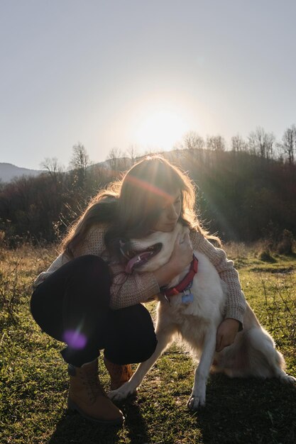 Foto jonge blanke vrouw met lang haar zit in het veld bij zonsondergang en knuffelt een witte bastaardhond