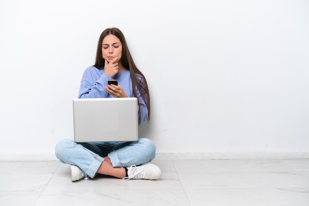 Jonge blanke vrouw met laptop zittend op de vloer geïsoleerd op een witte achtergrond denken en een bericht verzenden
