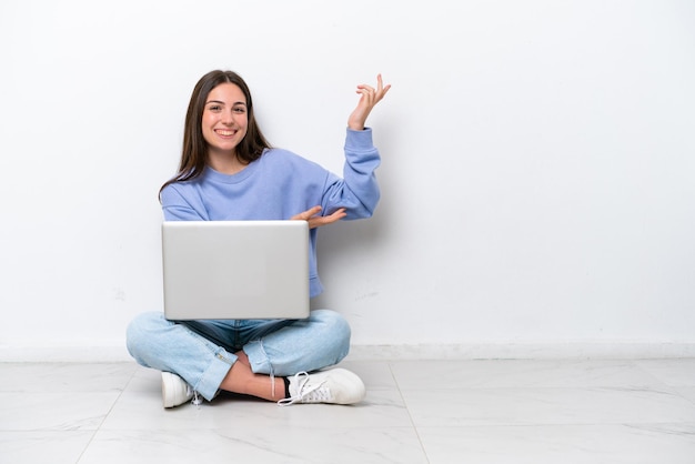 Jonge blanke vrouw met laptop zittend op de vloer geïsoleerd op een witte achtergrond die de handen naar de zijkant uitstrekt om uit te nodigen om te komen