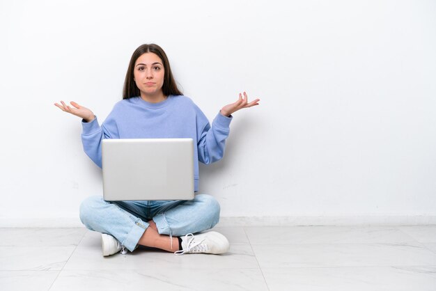 Jonge blanke vrouw met laptop zittend op de vloer geïsoleerd op een witte achtergrond die twijfels heeft terwijl ze haar handen opsteekt