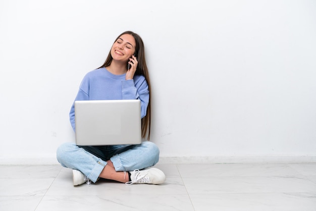 Jonge blanke vrouw met laptop zittend op de vloer geïsoleerd op een witte achtergrond een gesprek voeren met de mobiele telefoon