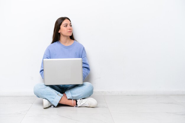 Jonge blanke vrouw met laptop zittend op de vloer geïsoleerd op een witte achtergrond in zijpositie