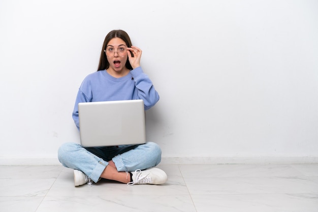 Jonge blanke vrouw met laptop zittend op de vloer geïsoleerd op een witte achtergrond met een bril en verrast