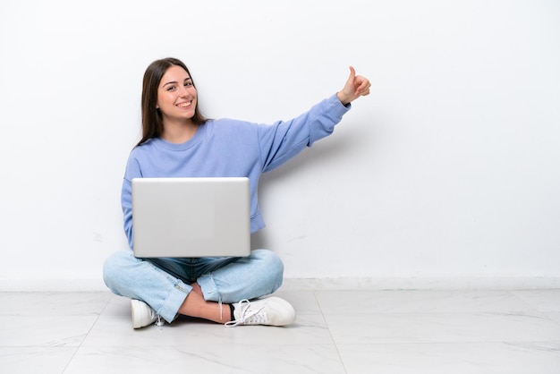 Jonge blanke vrouw met laptop zittend op de vloer geïsoleerd op een witte achtergrond met een duim omhoog gebaar