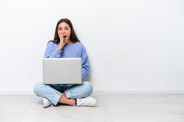 Jonge blanke vrouw met laptop zittend op de vloer geïsoleerd op een witte achtergrond met verrassing en geschokte gezichtsuitdrukking