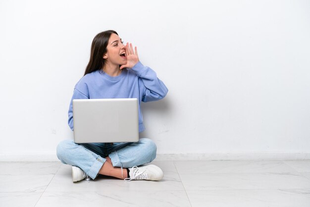 Jonge blanke vrouw met laptop zittend op de vloer geïsoleerd op een witte achtergrond schreeuwend met de mond wijd open naar de laterale