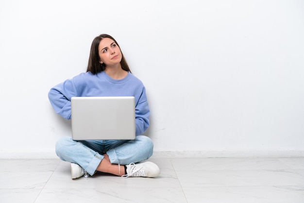 Jonge blanke vrouw met laptop zittend op de vloer geïsoleerd op een witte achtergrond twijfelend gebaar maken terwijl het opheffen van de schouders