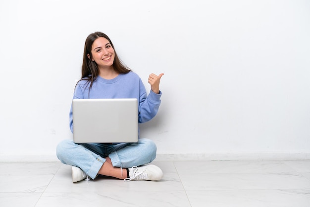 Jonge blanke vrouw met laptop zittend op de vloer geïsoleerd op een witte achtergrond wijzend naar de zijkant om een product te presenteren