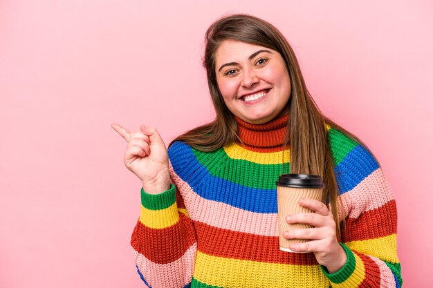 Jonge blanke vrouw met overgewicht die afhaalkoffie houdt geïsoleerd op een roze achtergrond glimlachend en opzij wijzend met iets op lege ruimte