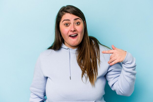 Jonge blanke vrouw met overgewicht geïsoleerd op een blauwe achtergrond lachend om iets dat de mond bedekt met handen