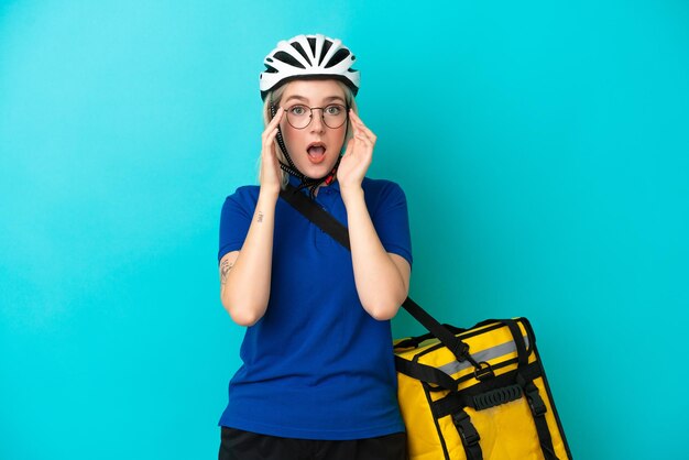 Jonge blanke vrouw met thermische rugzak geïsoleerd op blauwe achtergrond met een bril en verrast