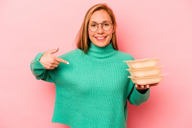 Jonge blanke vrouw met tupperware geïsoleerd op een roze achtergrond persoon die met de hand wijst naar een shirt kopieerruimte trots en zelfverzekerd?