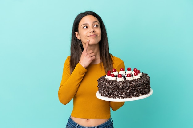 Jonge blanke vrouw met verjaardagstaart geïsoleerd op een blauwe achtergrond die twijfelt terwijl ze omhoog kijkt