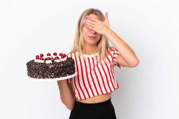 Jonge blanke vrouw met verjaardagstaart geïsoleerd op een witte achtergrond die ogen bedekt door handen