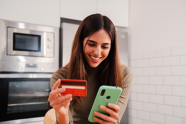 Jonge blanke vrouw met witte perfecte tanden glimlachend en een mobiele telefoon-app gebruiken om te kopen en te betalen met een creditcard om thuis te zitten keuken Vrouw geld uitgeven aan online winkelen op internet