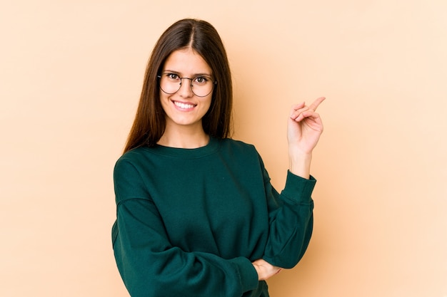 Jonge blanke vrouw op beige muur lacht vrolijk wijzend met wijsvinger weg.