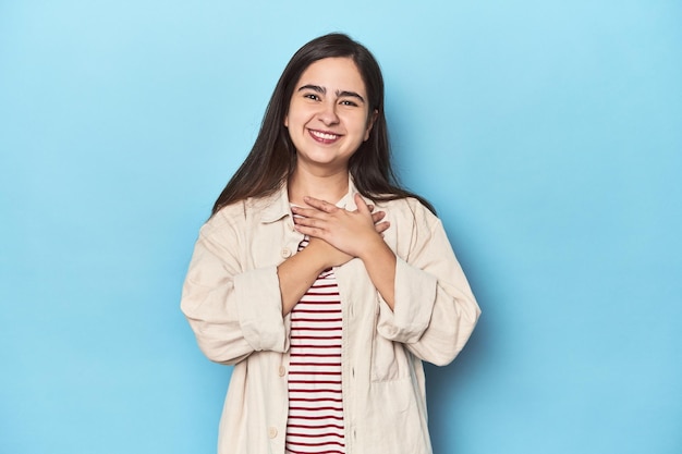 Jonge blanke vrouw op blauwe achtergrond heeft een vriendelijke uitdrukking en drukt de handpalm op de borst. Liefdesconcept