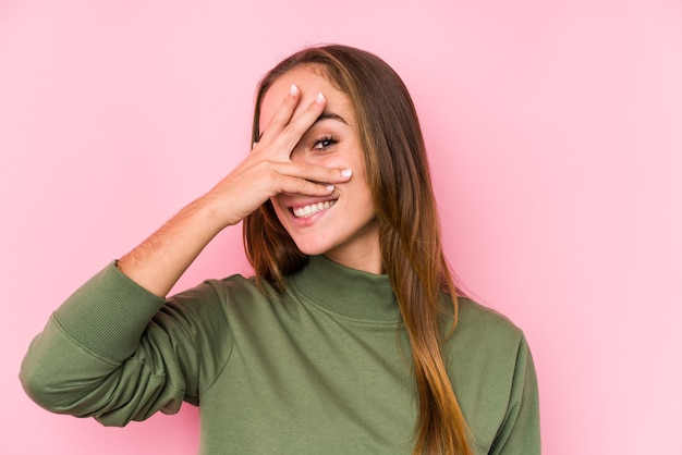 Jonge blanke vrouw poseren geïsoleerd knipperen naar de camera door vingers, beschaamd bedekkend gezicht.