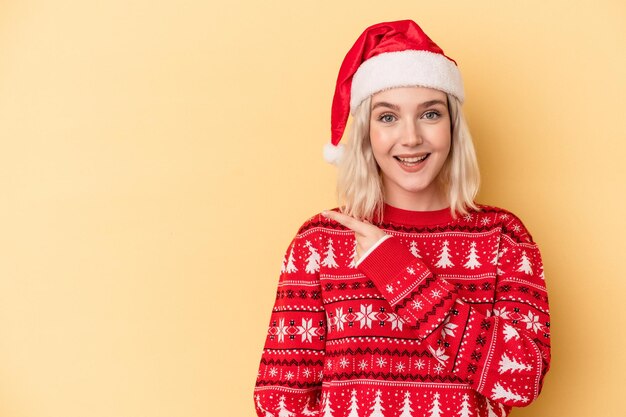 Jonge blanke vrouw viert Kerstmis geïsoleerd op gele achtergrond glimlachend en opzij wijzend, iets tonend op lege ruimte.