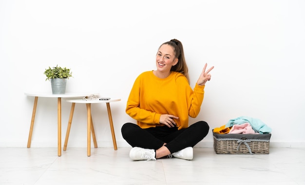 Jonge blanke vrouw vouwen kleren zittend op de vloer geïsoleerd op een witte achtergrond glimlachend en overwinning teken tonen