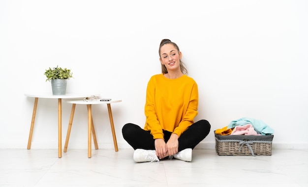 Jonge blanke vrouw vouwen kleren zittend op de vloer geïsoleerd op een witte achtergrond met gekruiste armen en happy