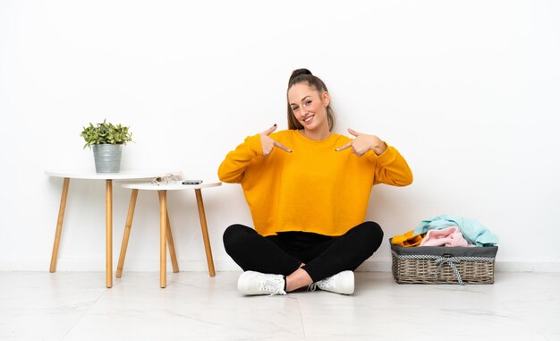 Jonge blanke vrouw vouwen kleren zittend op de vloer geïsoleerd op een witte achtergrond trots en zelfvoldaan