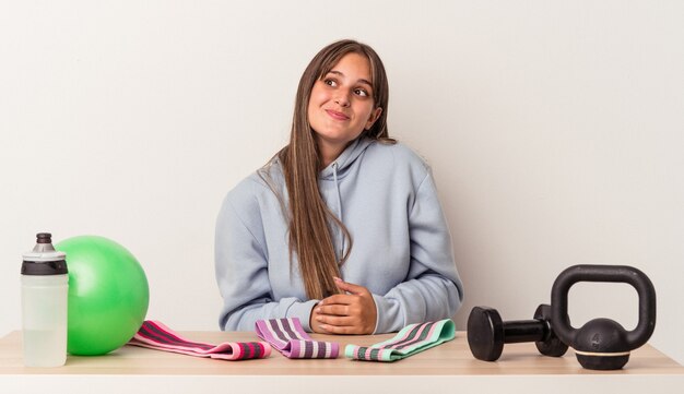 Foto jonge blanke vrouw zittend aan een tafel met sportuitrusting geïsoleerd op een witte achtergrond, dromend van het bereiken van doelen en doeleinden