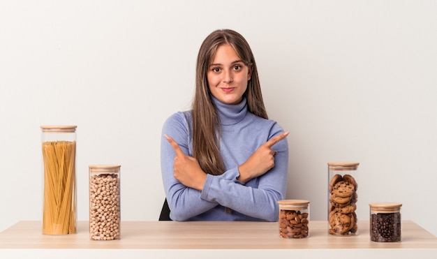 Jonge blanke vrouw zittend aan een tafel met voedselpot geïsoleerd op een witte achtergrond wijst zijwaarts, probeert te kiezen tussen twee opties.