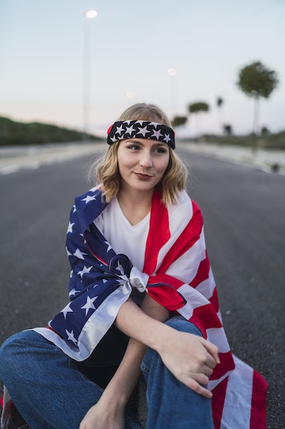 Jonge blanke vrouw zittend op de asfaltweg met de vlag van de vs op haar schouders