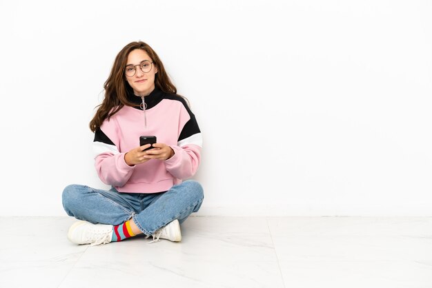 Jonge blanke vrouw zittend op de vloer geïsoleerd op een witte achtergrond die een bericht verzendt met de mobiel