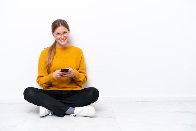 Jonge blanke vrouw zittend op de vloer geïsoleerd op een witte achtergrond die een bericht verzendt met de mobiel