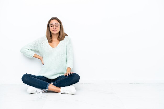 Jonge blanke vrouw zittend op de vloer geïsoleerd op een witte achtergrond die lijdt aan rugpijn omdat ze moeite heeft gedaan