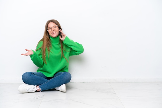 Jonge blanke vrouw zittend op de vloer geïsoleerd op een witte achtergrond een gesprek voeren met de mobiele telefoon met iemand