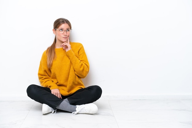 Jonge blanke vrouw zittend op de vloer geïsoleerd op een witte achtergrond en denkt aan een idee terwijl ze omhoog kijkt