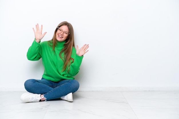 Jonge blanke vrouw zittend op de vloer geïsoleerd op een witte achtergrond en telt tien met vingers