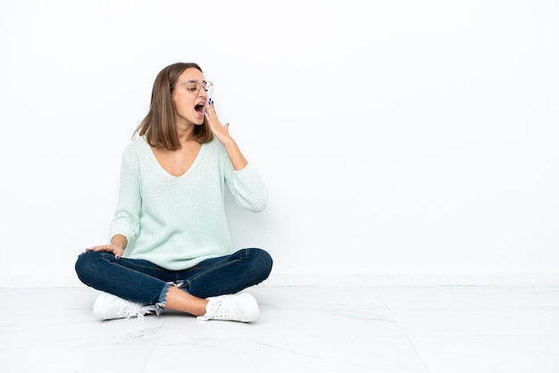 Jonge blanke vrouw zittend op de vloer geïsoleerd op een witte achtergrond geeuwen en wijd open mond met hand bedekken