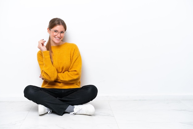 Jonge blanke vrouw zittend op de vloer geïsoleerd op een witte achtergrond met een bril en happy