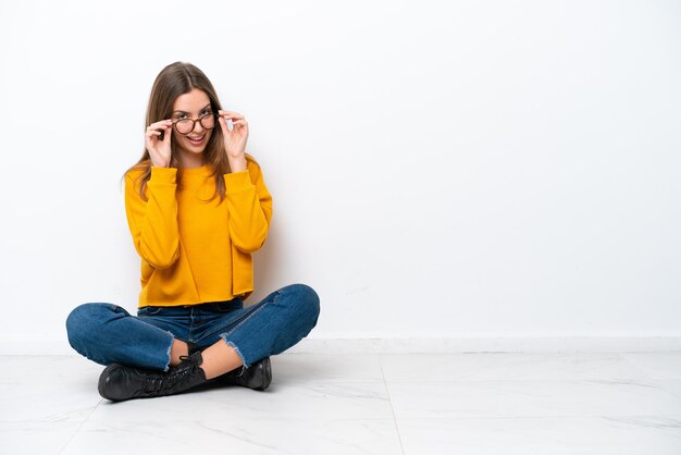 Jonge blanke vrouw zittend op de vloer geïsoleerd op een witte achtergrond met een bril en verrast