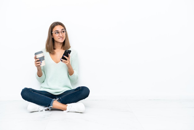 Jonge blanke vrouw zittend op de vloer geïsoleerd op een witte achtergrond met koffie om mee te nemen en een mobiel terwijl ze aan iets denkt