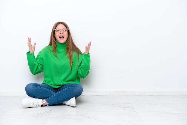 Jonge blanke vrouw zittend op de vloer geïsoleerd op een witte achtergrond met verrassing gezichtsuitdrukking