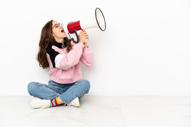 Jonge blanke vrouw zittend op de vloer geïsoleerd op een witte achtergrond schreeuwend door een megafoon