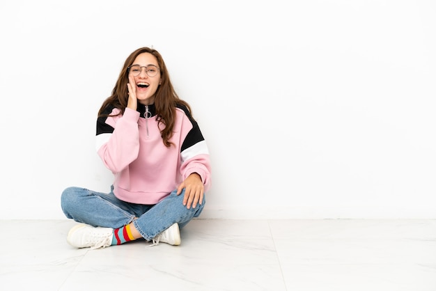 Jonge blanke vrouw zittend op de vloer geïsoleerd op een witte achtergrond schreeuwend met wijd open mond