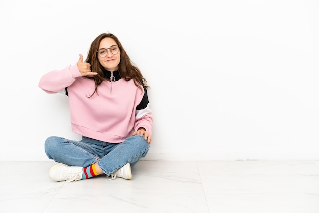 Jonge blanke vrouw zittend op de vloer geïsoleerd op een witte achtergrond telefoon gebaar maken. Bel me terug teken